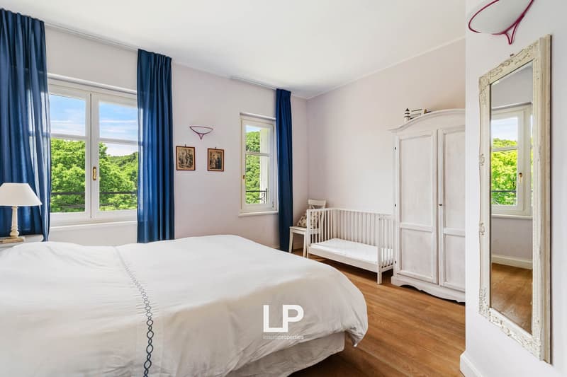 bedroom with white bed, wooden floor, blue curtains, white bedside lamp, white crib, white cabinet, mirror, white door, picture frames, small chandelier