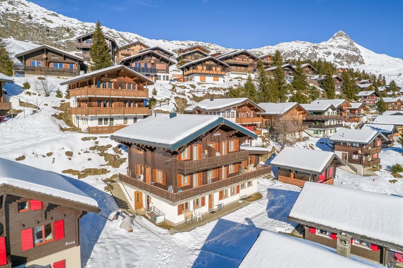 Mountain village, multiple wooden chalets, snow-covered rooftops, surrounded by trees and mountains