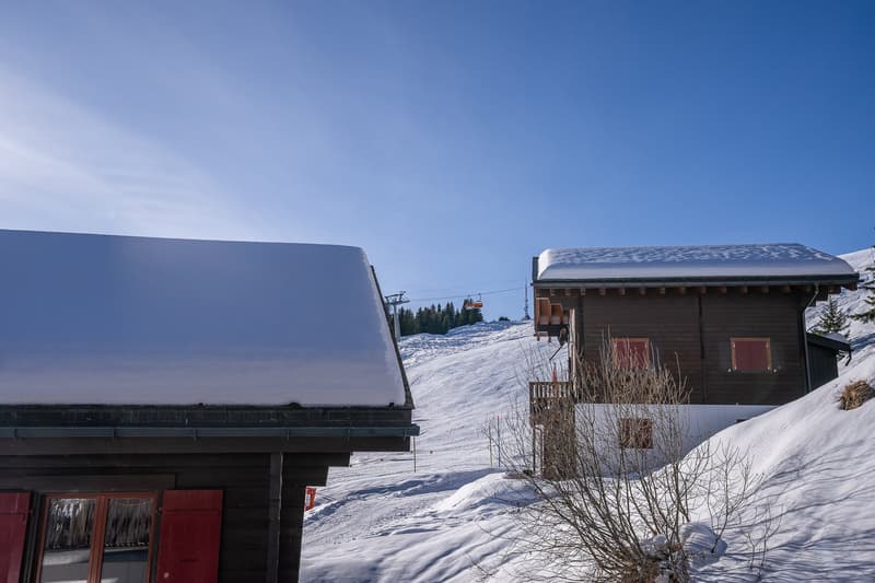 Two wooden chalets covered in snow, red shutters, wooden balconies, in a snowy landscape with ski lift