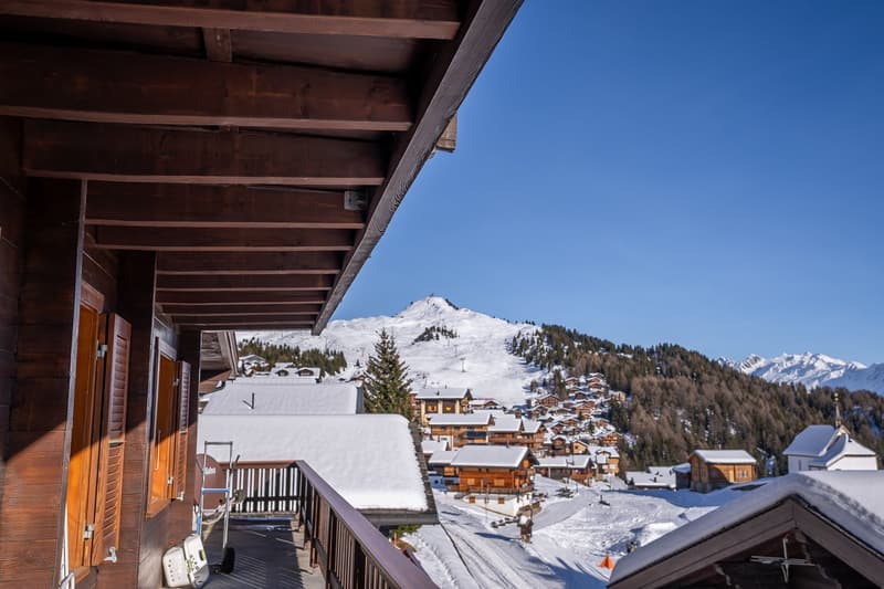 A balcony view from a building, overlooking a snowy landscape with mountains in the background. Wooden railings, a roof, and a wooden door.