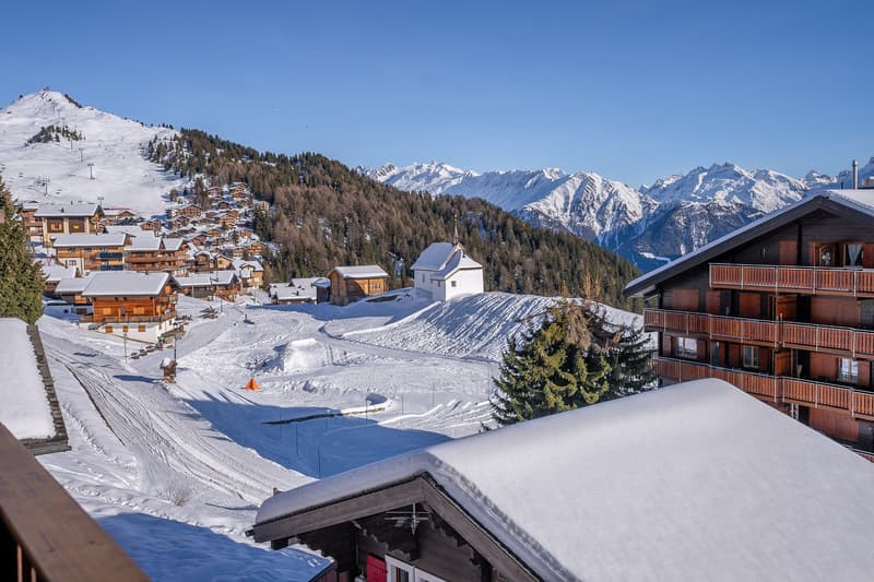 Mountain village with wooden chalets, snow-covered mountains, road, and a building with a balcony