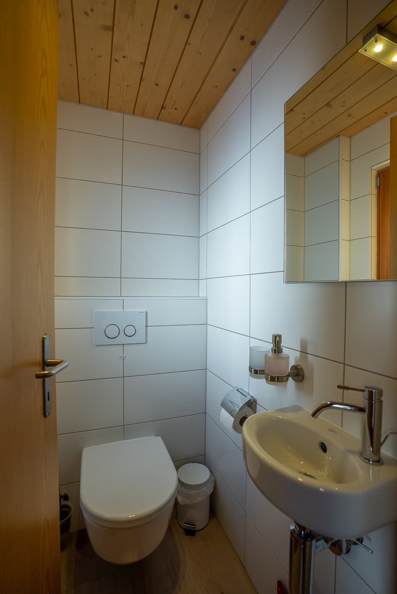 small room with white tiled walls, wooden floor, white toilet, sink, mirror