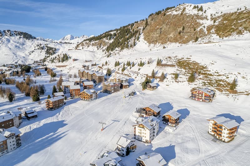 Aerial view of a ski resort with numerous chalets and lodges nestled amidst snow-covered mountains, a skiing slope, and surrounded by pine trees.