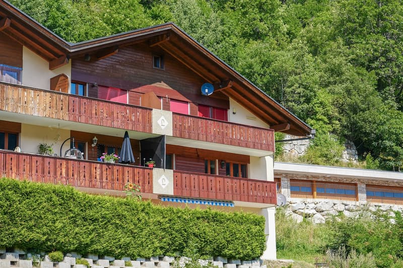 Apartment building, multiple wooden balconies, red shutters, hedge, grassy hillside, stone retaining wall