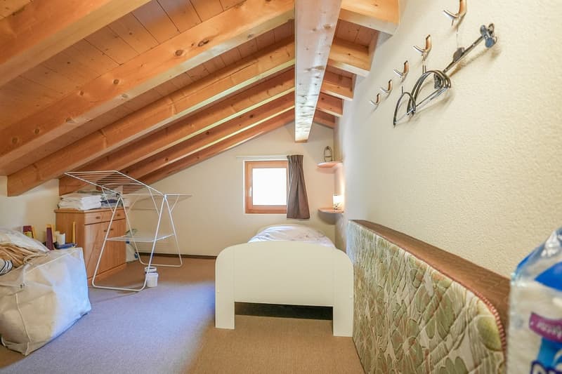 A room with wooden ceiling, small bed, wooden closet, window with curtain, carpeted floor, storage bag and drying rack.