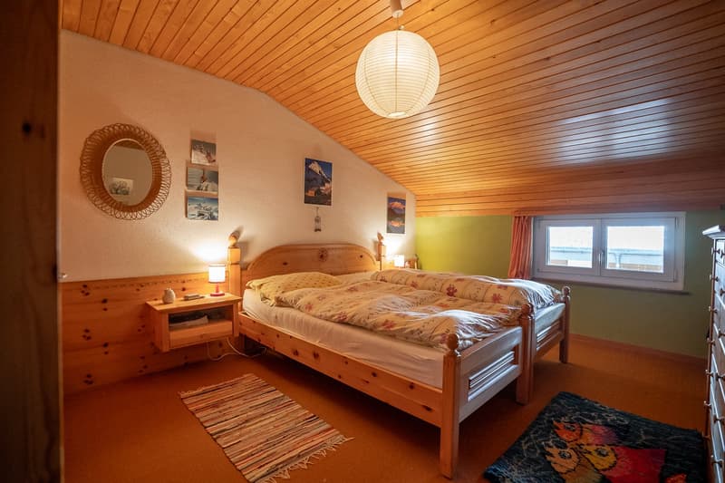 Bedroom with wooden beds, wooden ceiling, white walls, and wooden floor, two nightstands, curtains, framed pictures, and rugs.