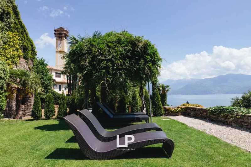 A garden with two lounge chairs on the grass near a walkway. Trees, bushes, and plants are around. There is a tower and a building on the left side, and a lake and mountains in the distance.