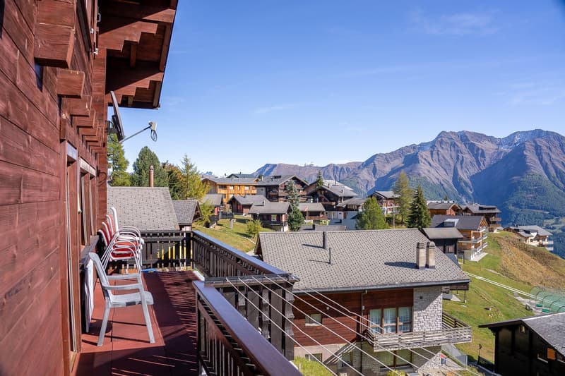 Balcony with chairs, wooden railings, surrounded by houses, mountainous landscape, clear blue sky