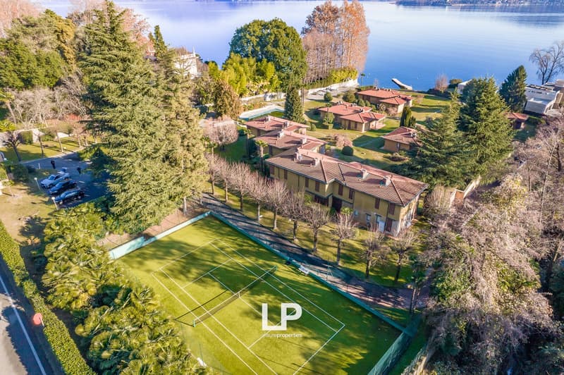 Several houses surrounded by trees and a tennis court, overlooking a lake.