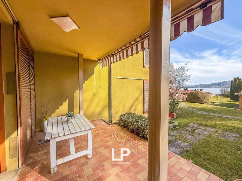 Outdoor space with tiled flooring, a table with a plant, yellow wall, an awning, a drain on the floor, a green garden, and a view of the lake in the distance.