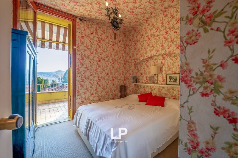 Double bed with white cover, red pillows, floral wallpaper, lamp, entrance with door, balcony