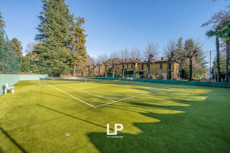outdoor tennis court, yellow house in the background, trees, bench, green fence