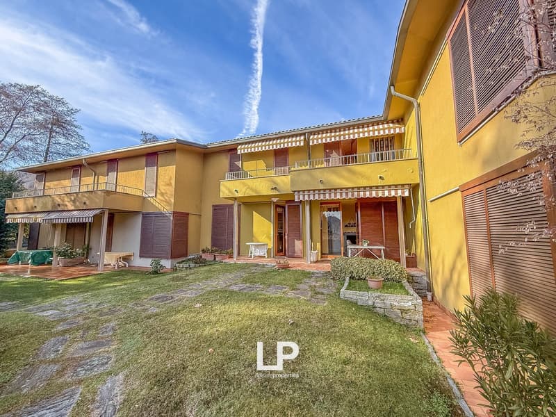 A yellow house with 2 floors, wooden window shutters, balconies with awnings, grassy yard, tables, plants