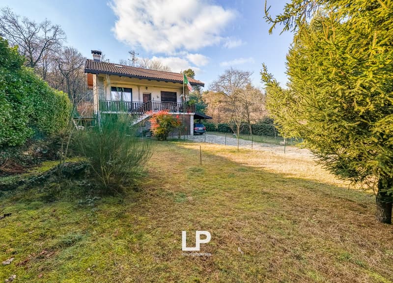 Two-story house, tile roof, balcony, driveway, car parked, grassy yard, shrubs, trees, flag on pole