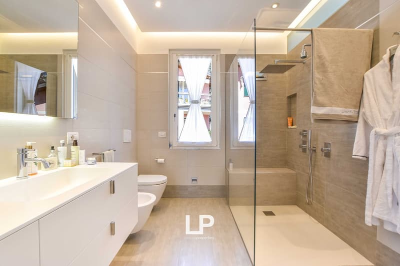 modern bathroom with white cabinets, sink, modern shower, large mirror, window with curtains, white floor tiles, toilet, bidet, robes and towel bar