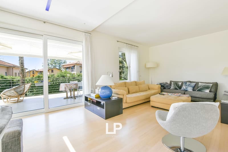 Spacious living room with large windows, hardwood floors, yellow sofa, gray armchair, coffee table, and access to a balcony or terrace overlooking a garden area