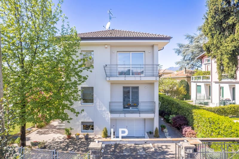 2-story white house with a tiled roof, balcony, and garage. The house is surrounded by trees and landscaping, with a paved driveway leading to the garage.