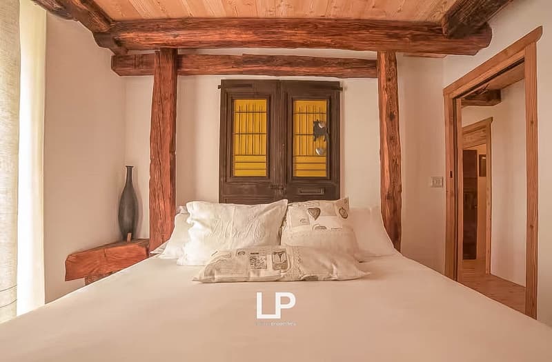Bedroom with wooden beams, large bed, pillows, nightstand with a vase, two double doors with yellow glass panels, wooden door.