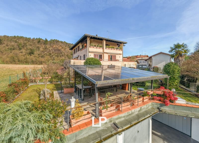 Two-story house with terrace and solar panels, surrounded by a well-maintained garden, featuring various plants and trees, and a paved driveway leading to the entrance.