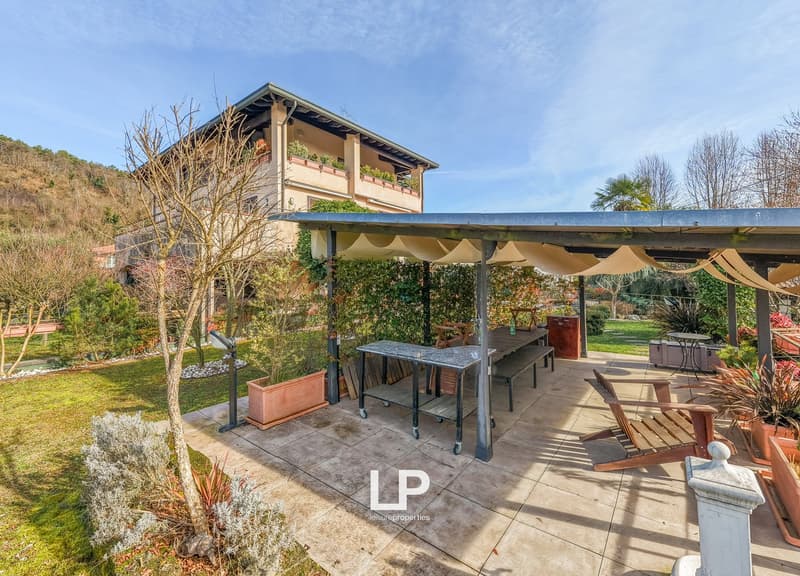 Outdoor space with a canopy, wooden benches, tables, and plants, overlooking a building