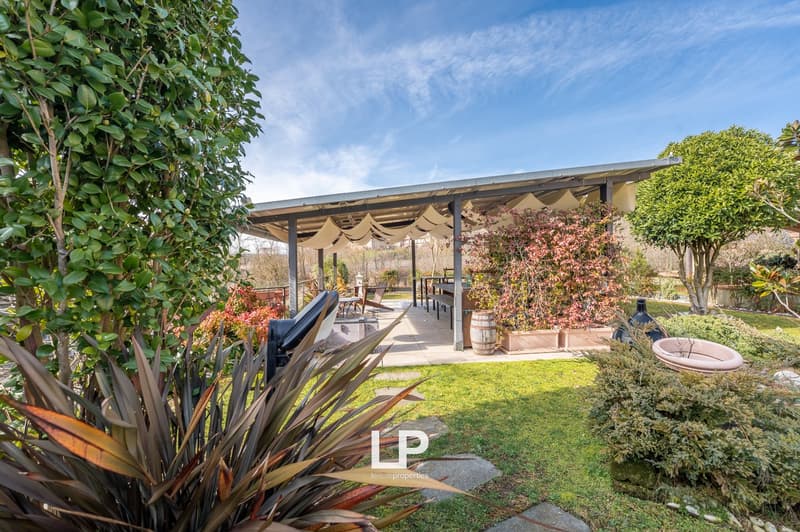 Garden with outdoor dining area, wooden pavilion, trees, shrubs, pathway, and clear sky.