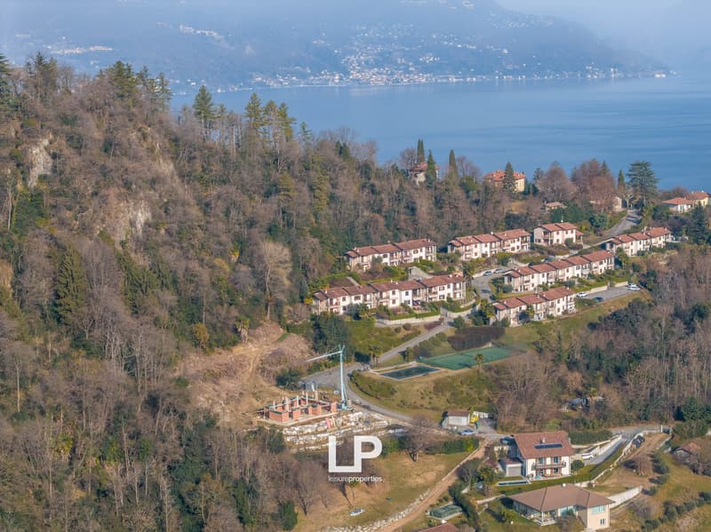 Aerial view of a hillside with several villas, surrounded by trees, a tennis court, and a swimming pool. There are mountain and sea views in the distance.