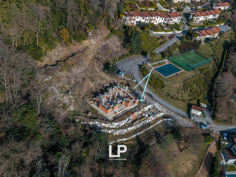 House under construction, tennis court, swimming pool, nearby road, surrounding trees, multiple buildings in the distance