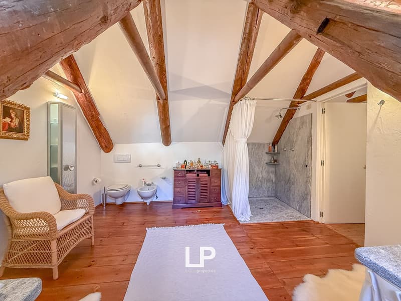 Wooden ceiling beams, wicker armchair, white rug, white porcelain bathroom sink, wooden cabinet, shower stall