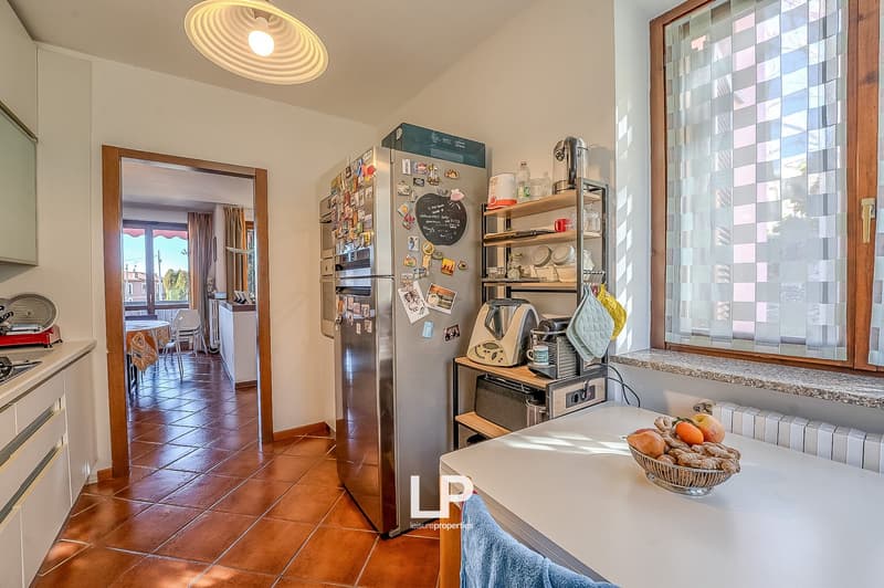 Stainless steel refrigerator, wooden shelves, gas stove, wooden table with a bowl of fruits, wooden flooring, white cabinets, white tiled floor