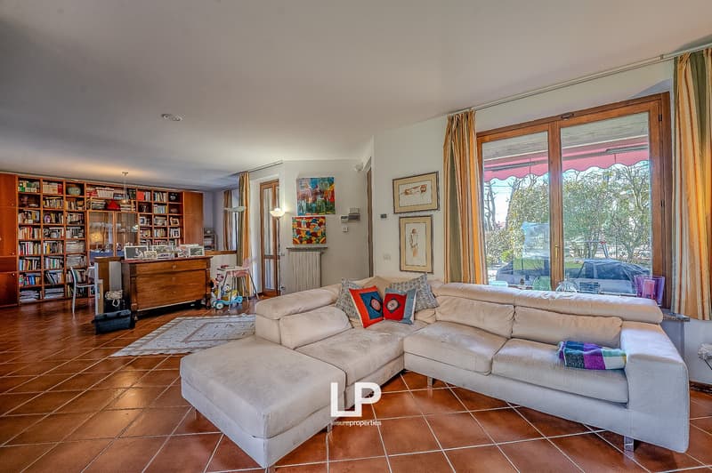 Living room with wooden floor, beige sofa, large window, decorative items on the wall, bookshelves, radiator, rug.