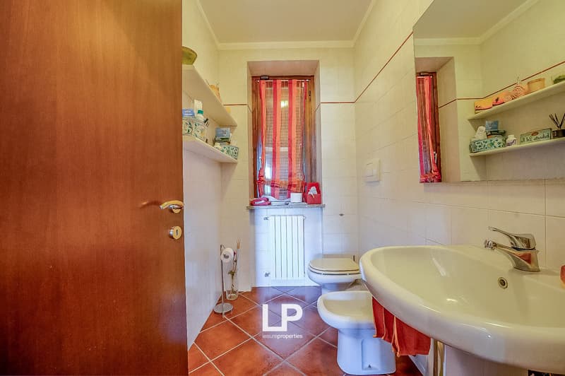 bathroom, white walls, white tile flooring, white sink, white bidet, toilet, radiator, window with red curtains, shelves