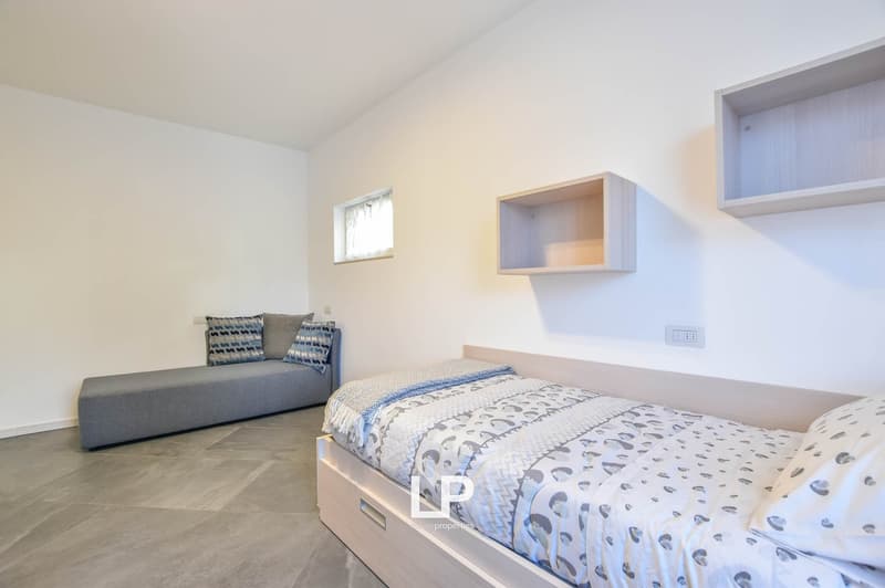 A single bed and a gray sofa with pillows, two small shelves on the wall, gray floor, white walls, white ceiling, a power outlet, and a small window with white curtains.