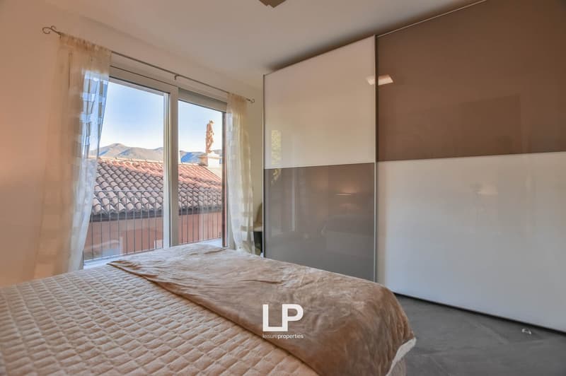 Bedroom with large bed, white walls, glass windows with curtains, balcony, large white and brown closet.