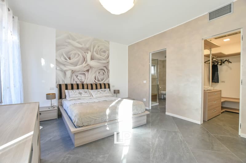 Double bed, wooden headboard, two nightstands with lamps, white walls, decorative wallpaper, gray tiled floor, large window with curtains, wardrobe with clothes hanger