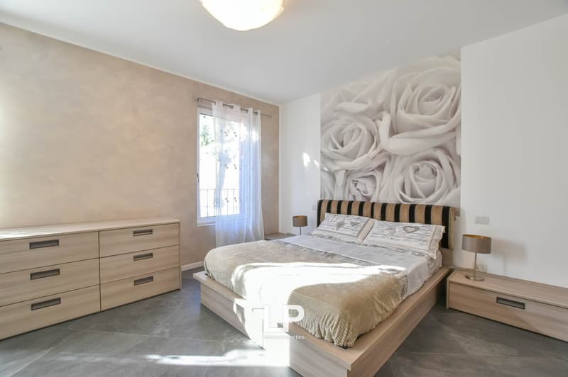 spacious room, large bed, wooden bed frame, bedspread, two pillows, two nightstands with lamps, wooden dresser with six drawers, large mural of white roses on the wall, window with white curtains, tiled floor