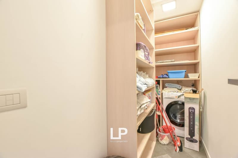 A room with built-in shelves and a washing machine in the corner, a broom, and a bucket beside the washing machine, an open box next to the washing machine, and a light on the ceiling.