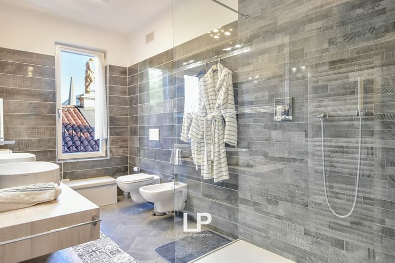 Modern bathroom with glass walls, white porcelain sink, two white toilets, shower with silver fixtures, towel hanging on a hook, and decorative tiles