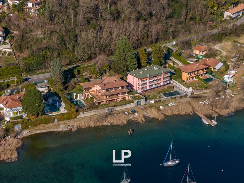 Aerial view of a residential area with several houses, swimming pool, and a waterfront. The houses are surrounded by trees and greenery. The waterfront has a dock with boats and a clear view of the water.