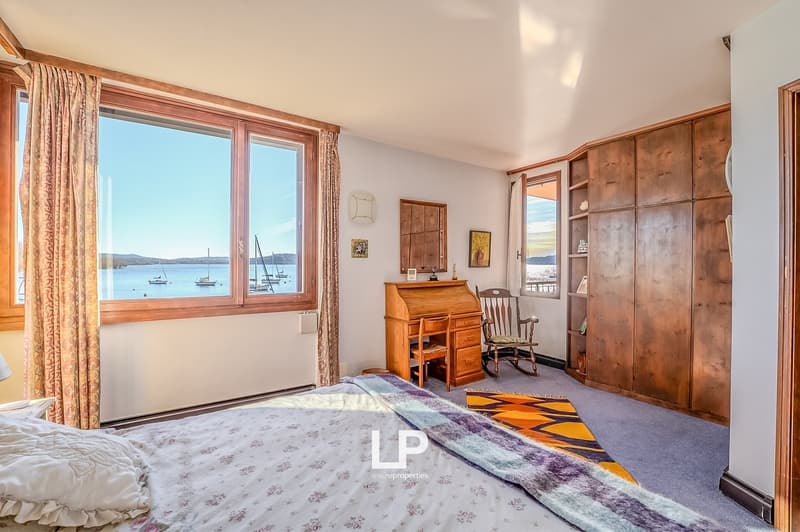bedroom with bed, desk, rocking chair, wooden cabinets, large windows with view of water and boats