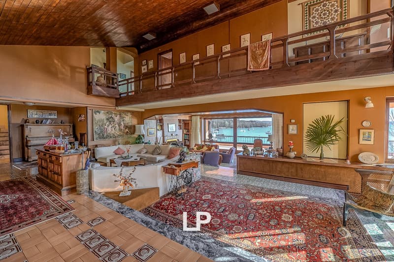 large living room, wooden ceiling, large windows, balcony, couches, wooden desk, rugs, piano, paintings, lamps