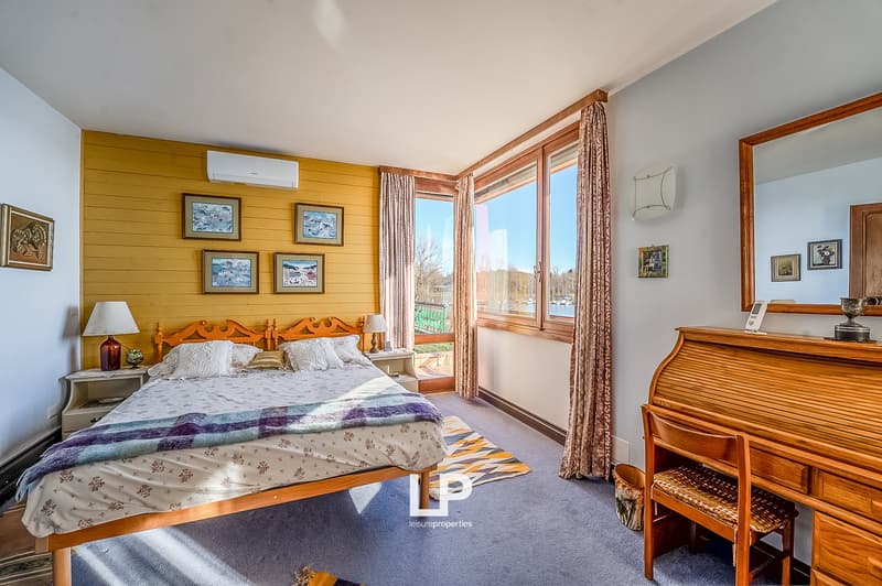 Bedroom with wooden bed, nightstands, lamps, air conditioner, large windows, wooden desk, mirror, rug, wooden chair, and picture frames on the wall.