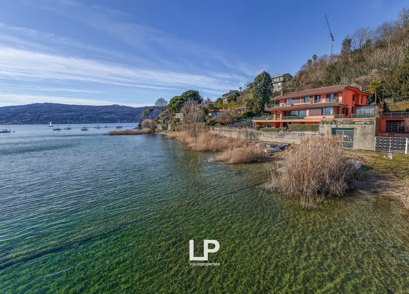 House on the lake shore, red exterior, multiple windows, surrounded by greenery, mountains in the background