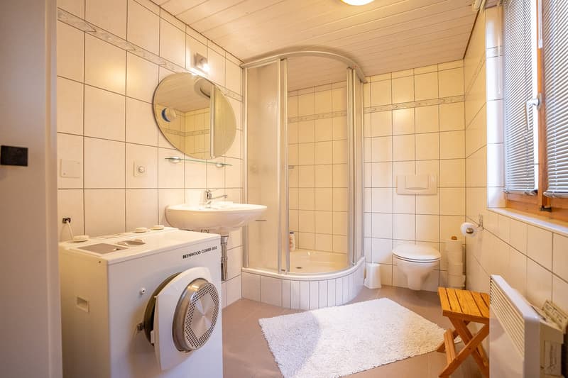 white tiled walls, white sink, shower, toilet, washing machine, wooden stool, white rug