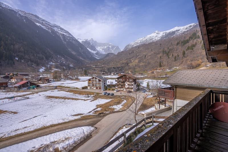 Mountain view, wooden balcony, snow-covered valley, several buildings, trees, mountains