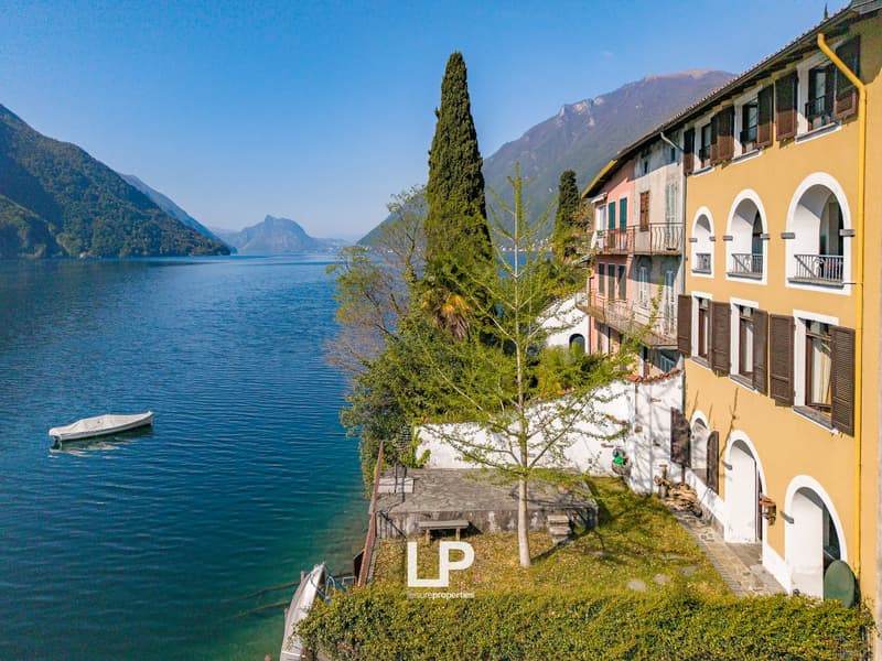 House on the lakefront, yellow and pink facades, balcony, small boat, trees, mountains in the background