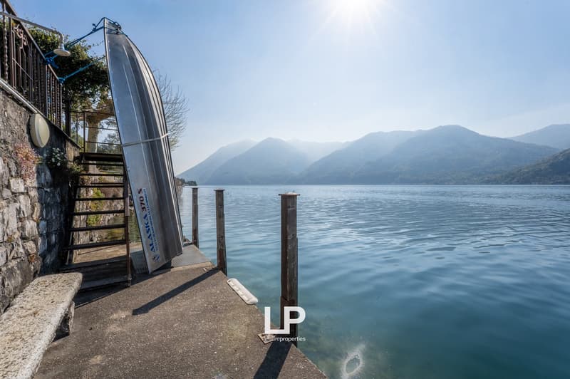 A boat is docked on a pier with a stone wall, a staircase, and a lamp. The pier extends into a body of water with mountains in the background.