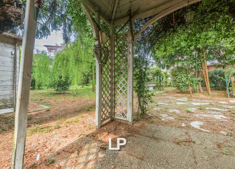 Garden with gravel path, wooden pergola, plants, trees, and a small building