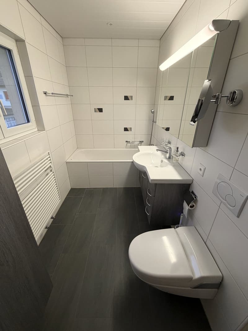 modern bathroom with white tiles, bathtub, sink, toilet, mirror, and a window