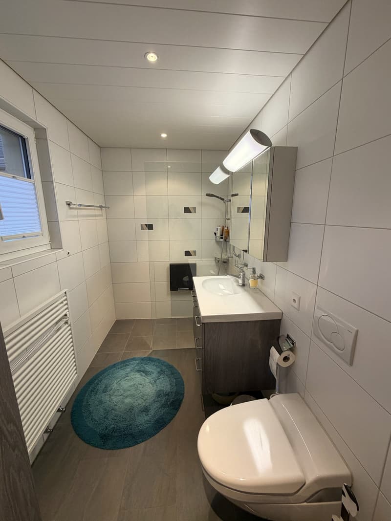 Bathroom with white tiles, white sink, mirror, toilet, blue rug, window, radiator, and shower