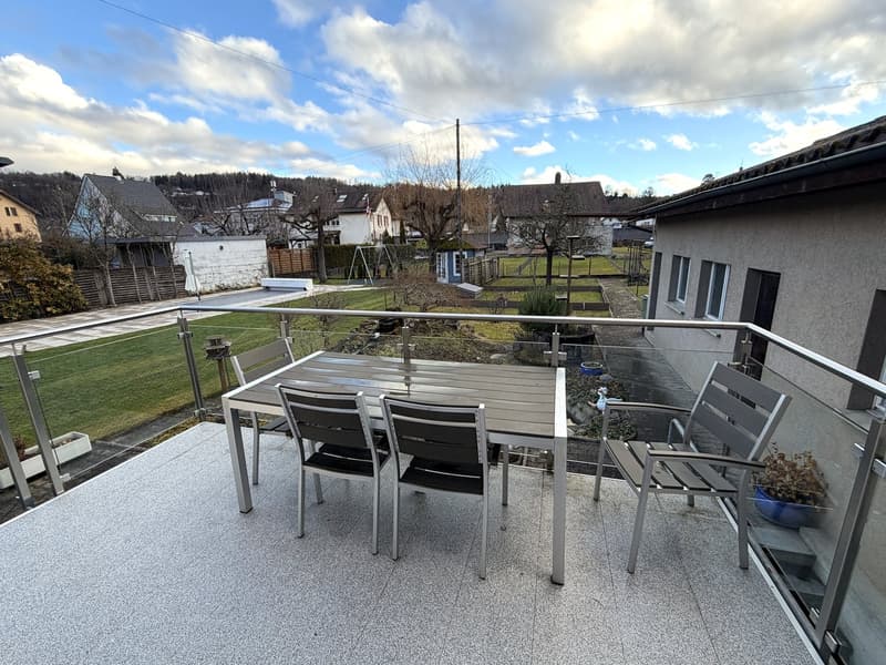Balcony with table and chairs, glass railing, potted plant, view of a grassy area and houses